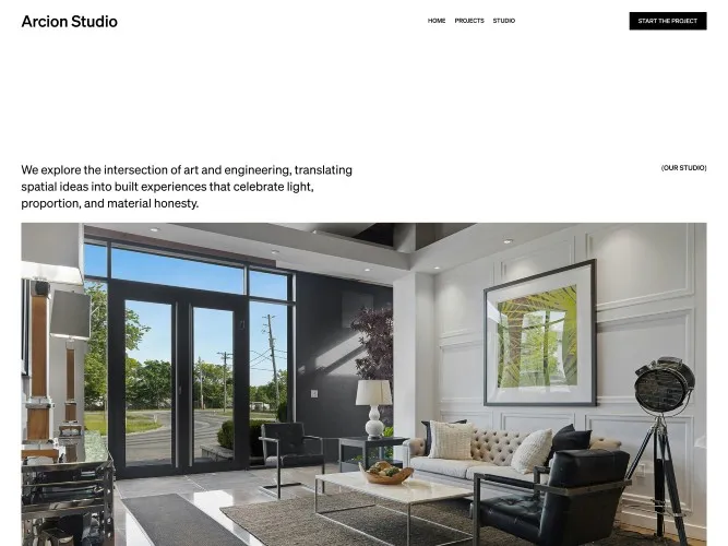 Modern studio living area with a beige tufted sofa, two black leather chairs, a marble coffee table, large framed artwork, and tall windows with a view of trees outside.