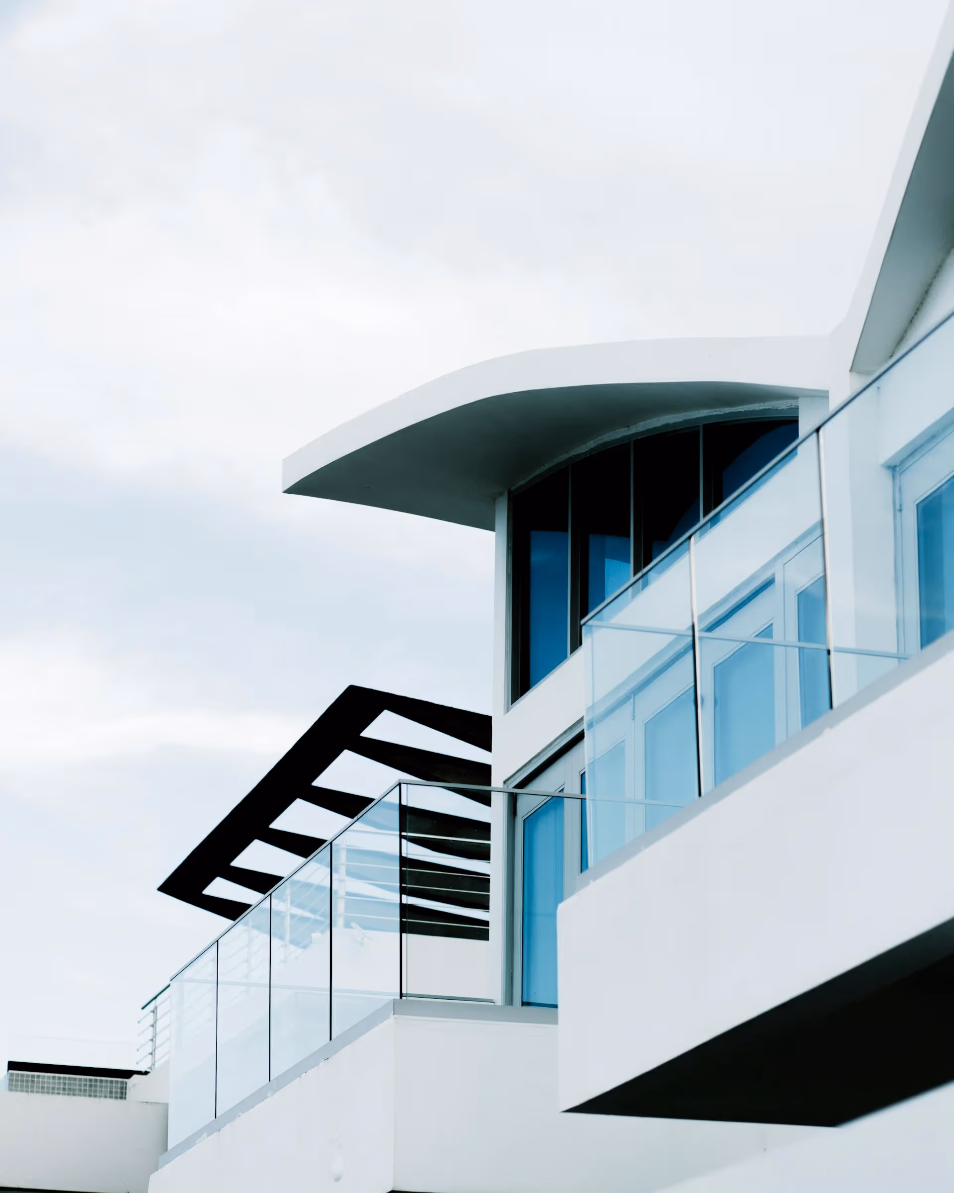 Modern white building with curved roof, large windows, and glass balcony railings under a cloudy sky.