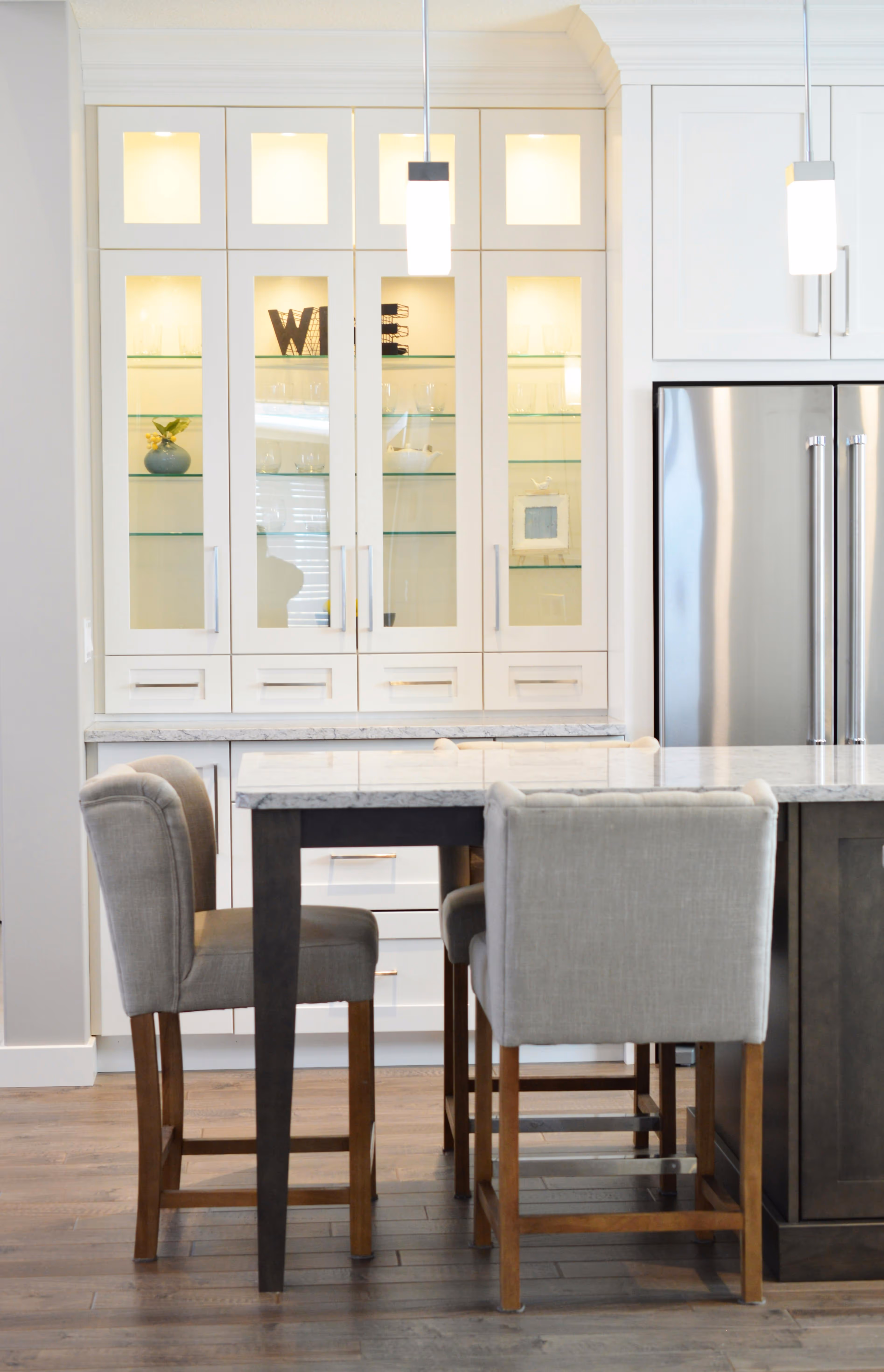 Modern kitchen with marble countertop island and four upholstered bar stools, white cabinetry with glass doors, and stainless steel refrigerator.